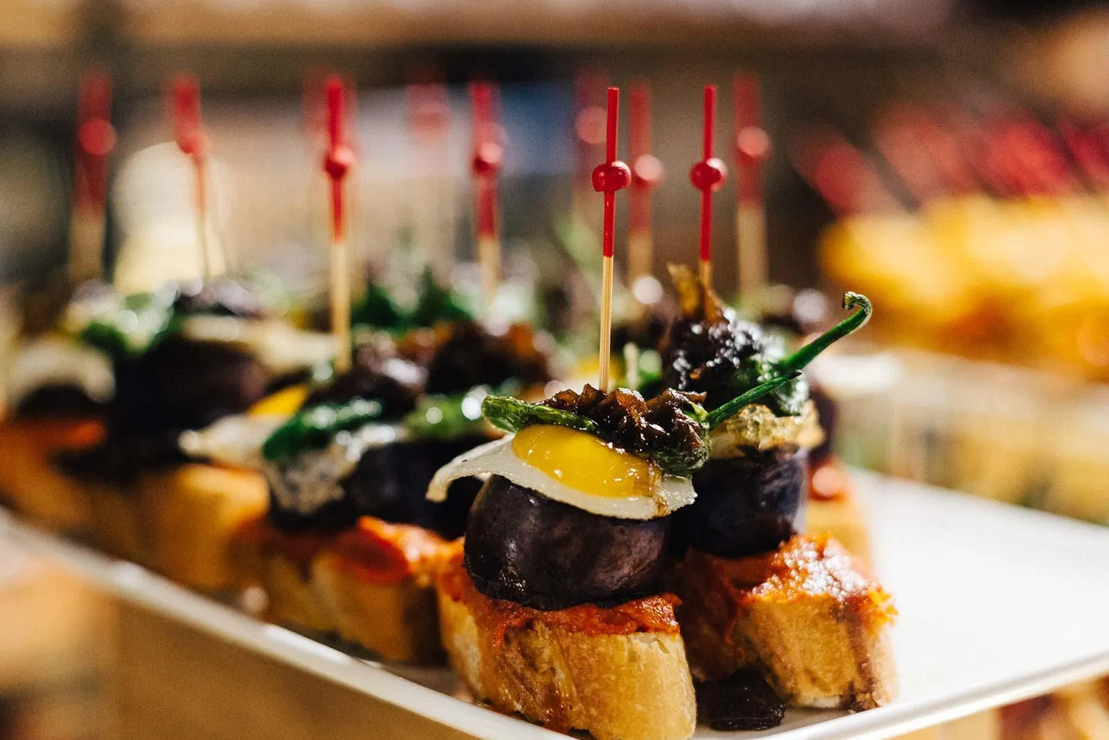 Nahaufnahme eines Tellers Pintxos, kleiner offener Sandwiches mit verschiedenen Belägen, gehalten von roten Zahnstochern. ...
