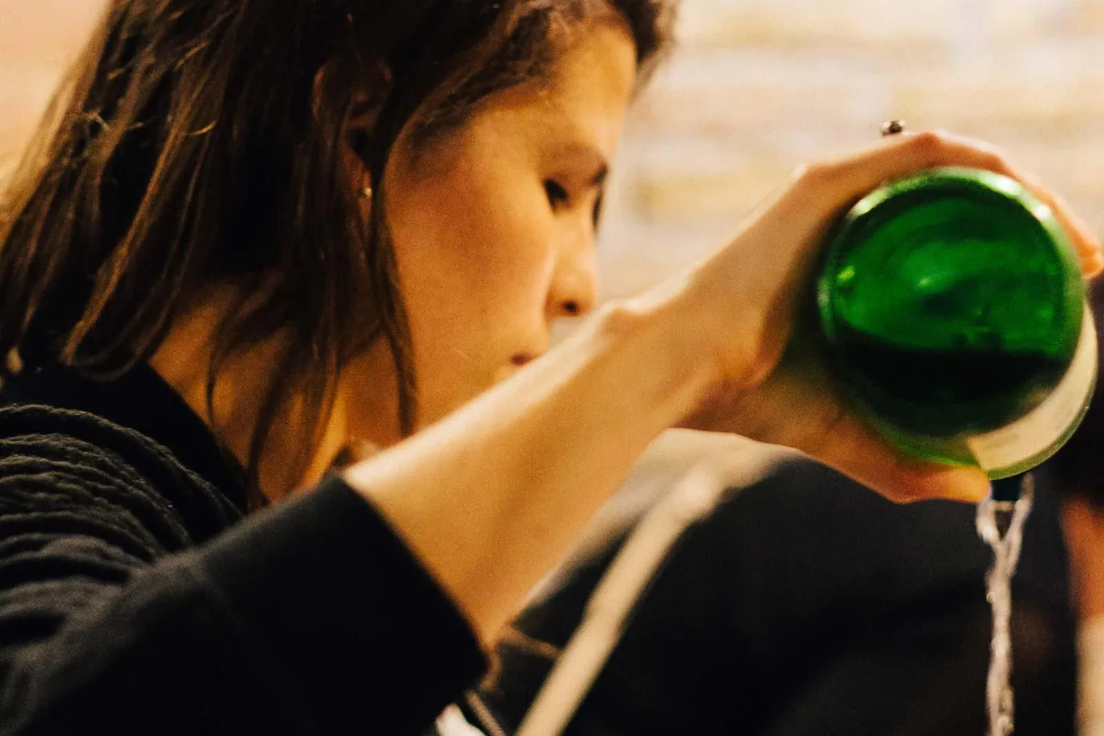 Person gießt Sidra aus grüner Flasche in Glas, Schaum erzeugend. Verschwommene Steinwand im Hintergrund.