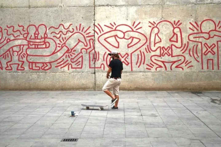 Person with skateboard in front of a concrete wall with red Keith Haring-style figures. A small blue object is on the ground.