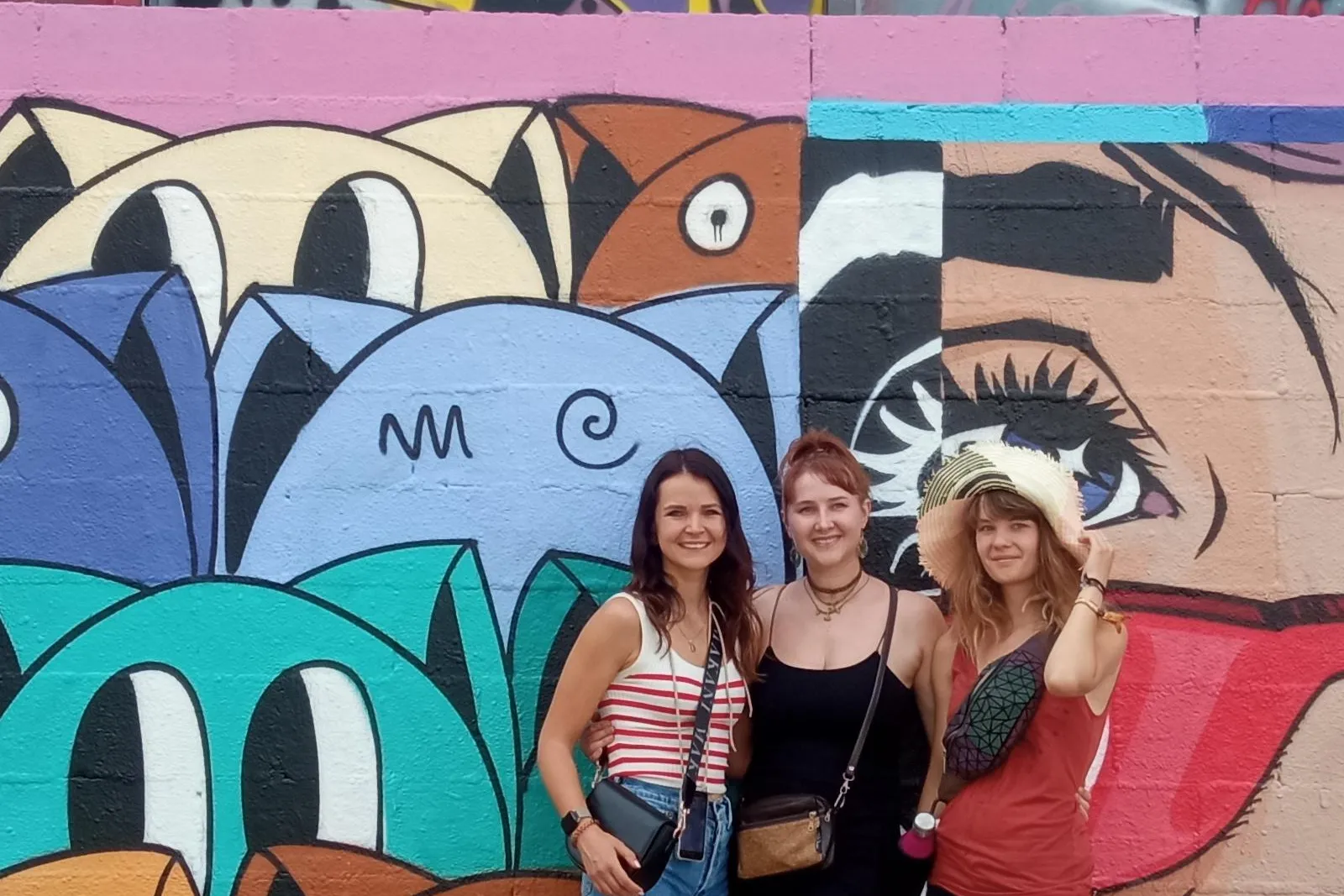 Drei lächelnde Frauen stehen vor einer farbenfrohen Wand mit abstraktem Graffiti und einem großen Wandgemälde mit stilisie...