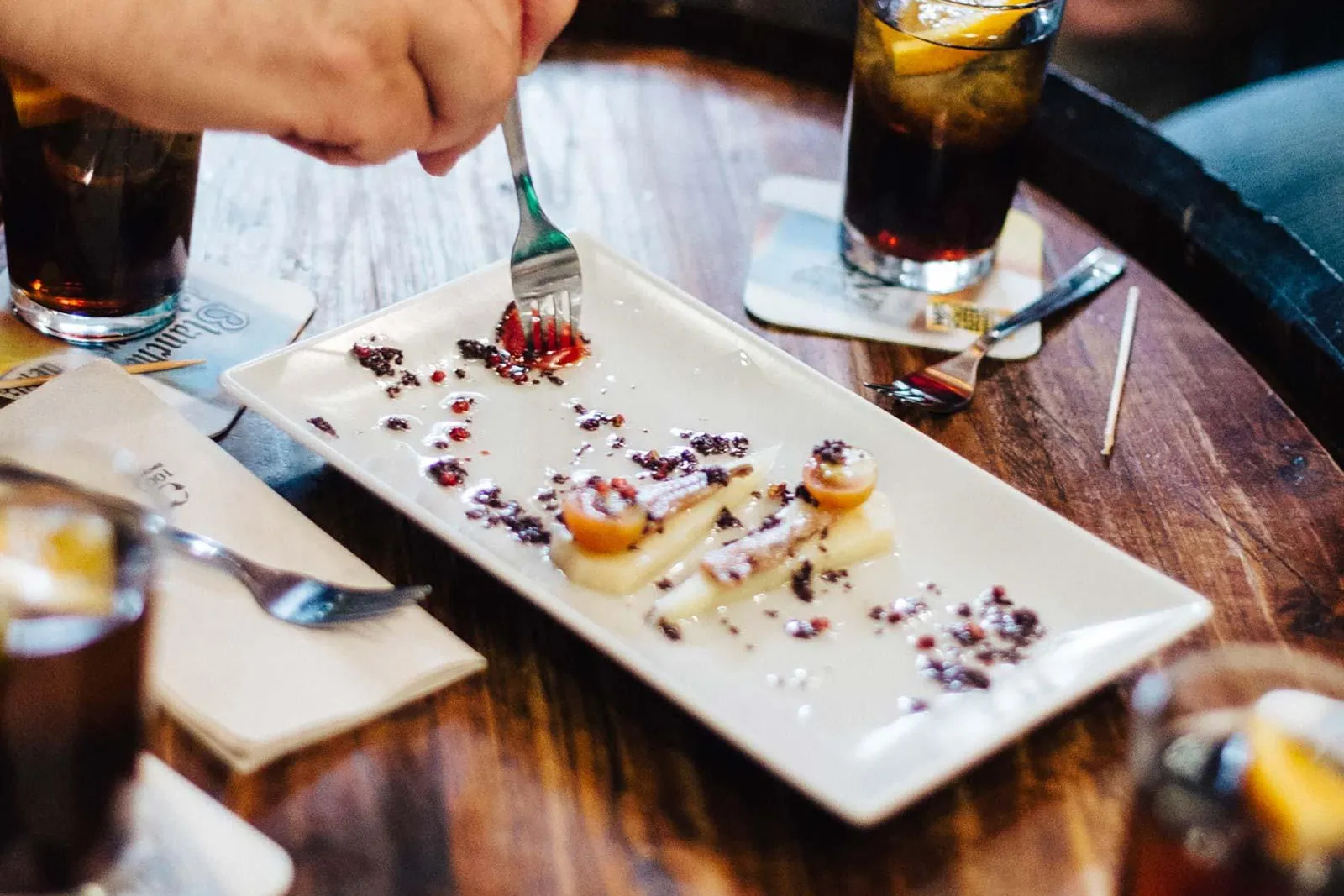 Hände von Personen an einem Holztisch mit Tapas und Getränken. Eine Person isst ein Gericht mit einer Gabel.