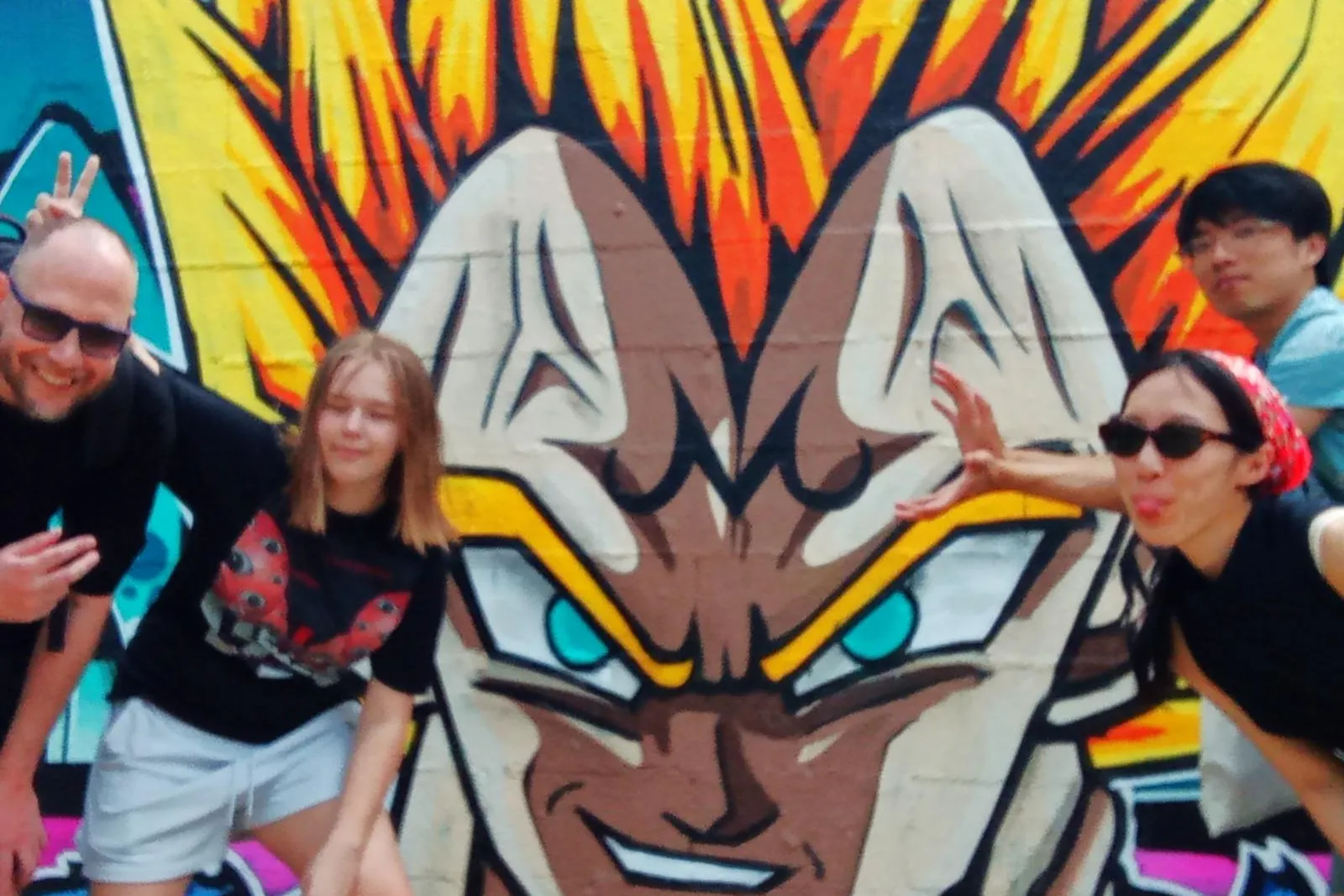 Four people posing in front of a colorful graffiti mural of a character with spiky yellow hair and blue eyes, under a clea...