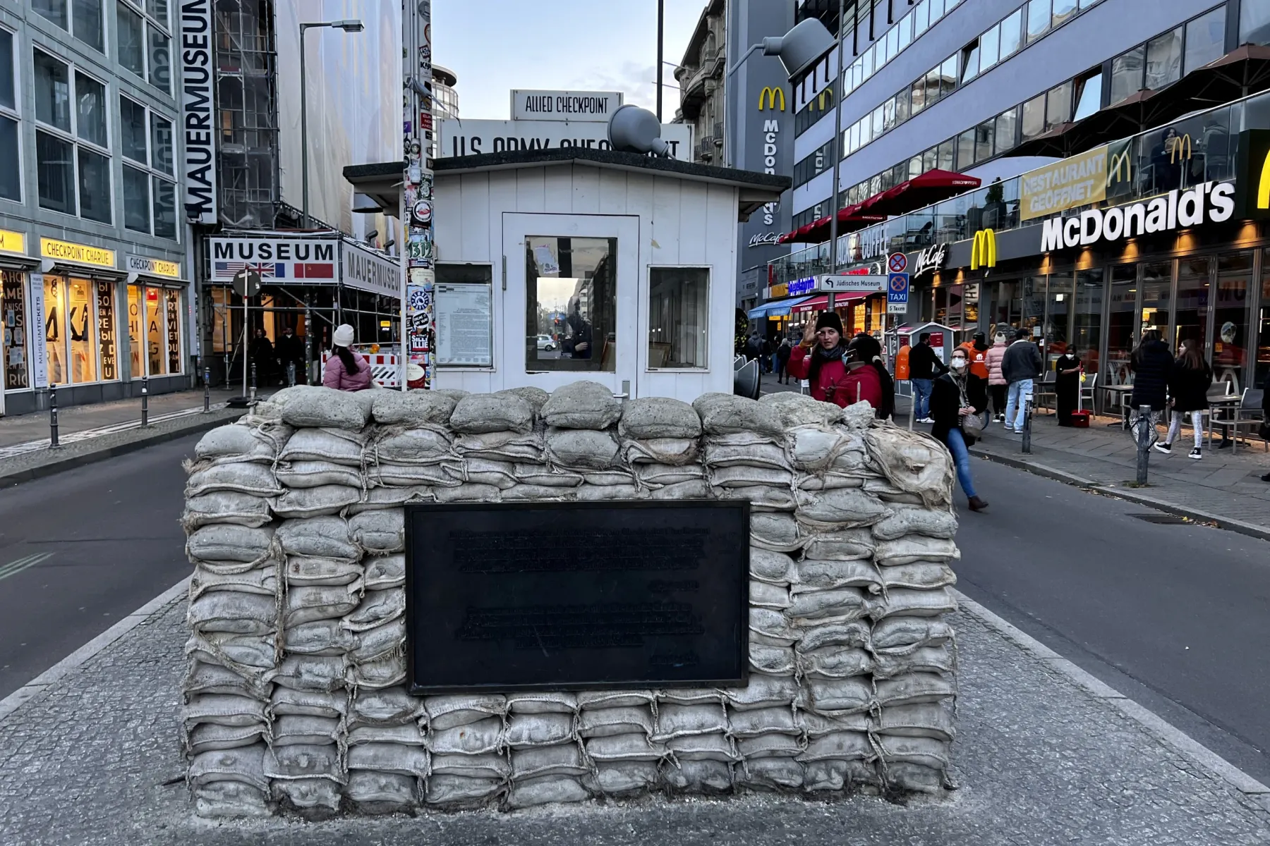 Reconstruction of Checkpoint Charlie guardhouse with sandbags and a plaque on a cobblestone street in Berlin, with buildin...