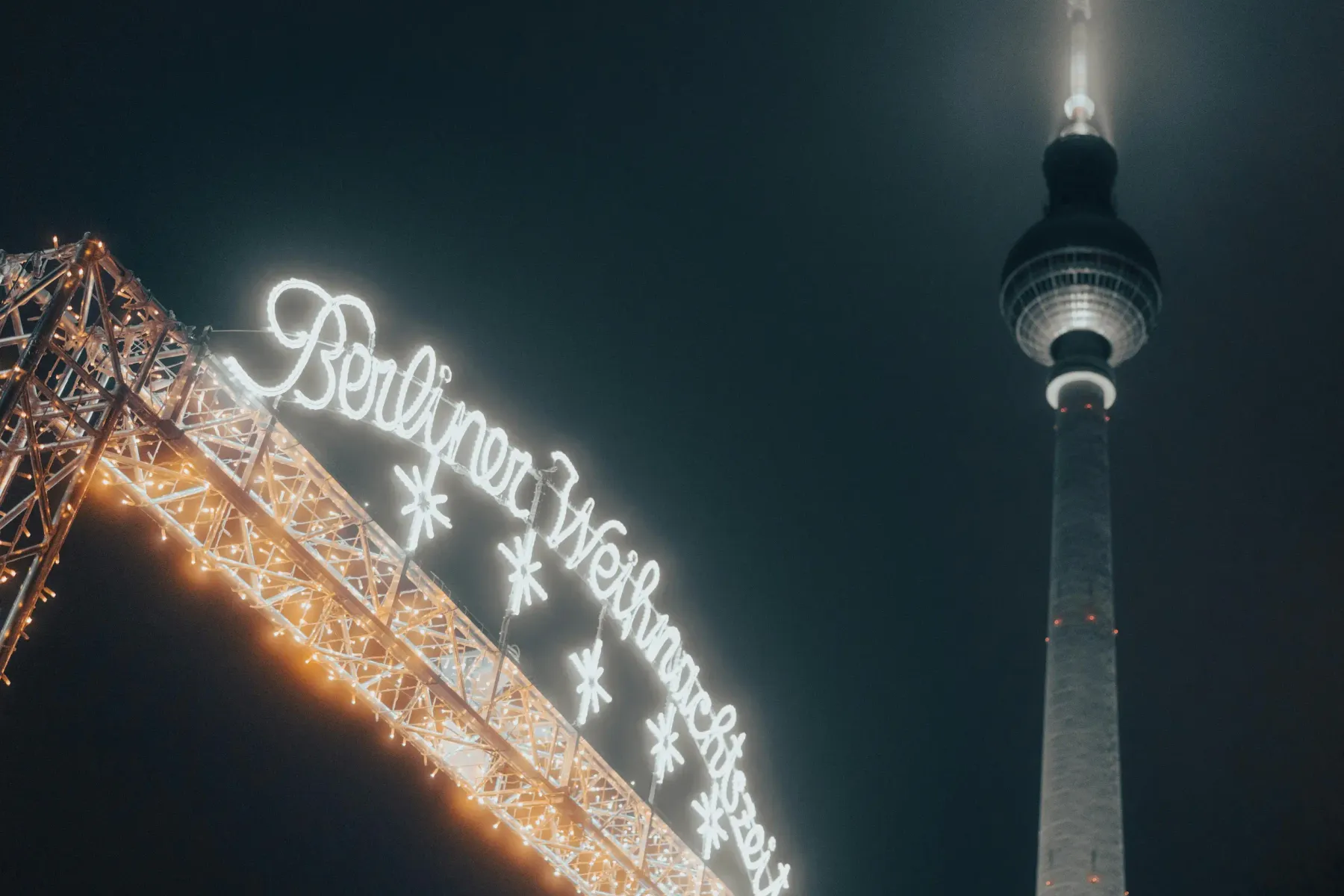Beleuchtetes Schild 'Berliner Weihnachtszeit' und ein Teil des Berliner Fernsehturms vor dunklem Nachthimmel.