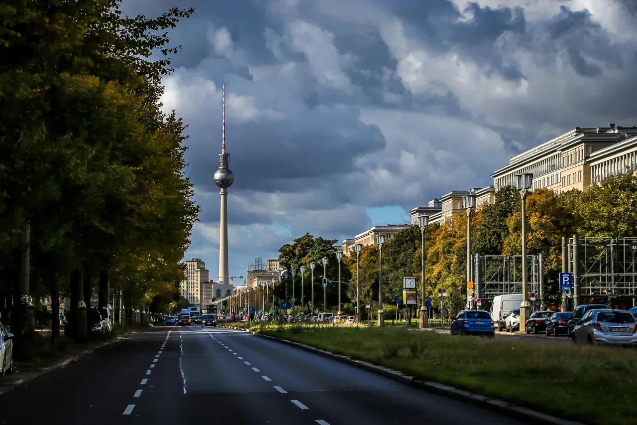 Vista de calle de la Torre de Televisión de Berlín (Fernsehturm) en un día nublado, con coches y árboles otoñales.