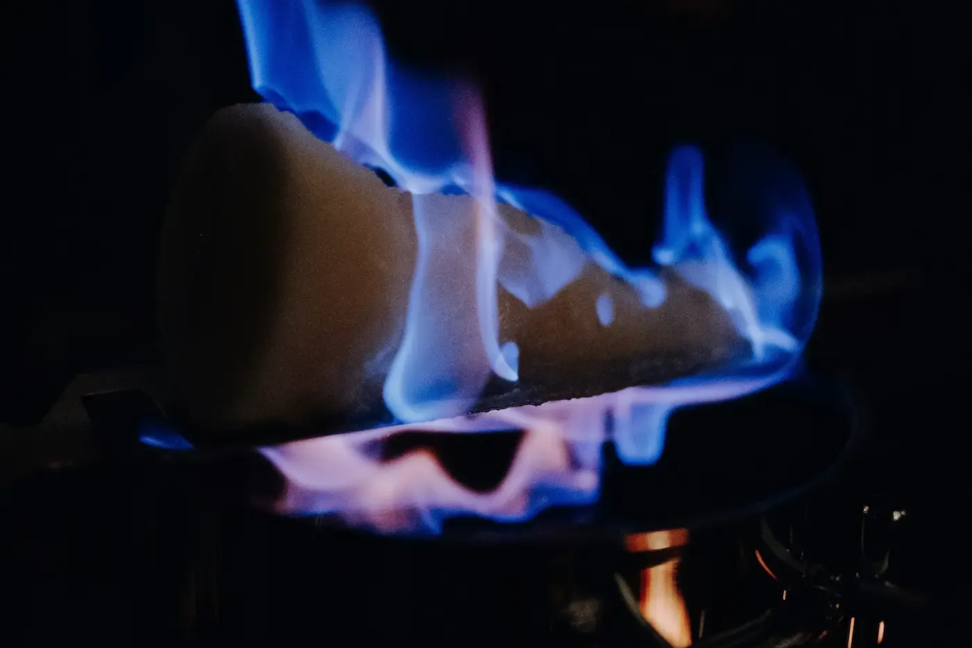 Zuckerhut mit blauen Flammen brennt über einem dunklen Topf, der den Vordergrund vor einem dunklen Hintergrund beleuchtet.