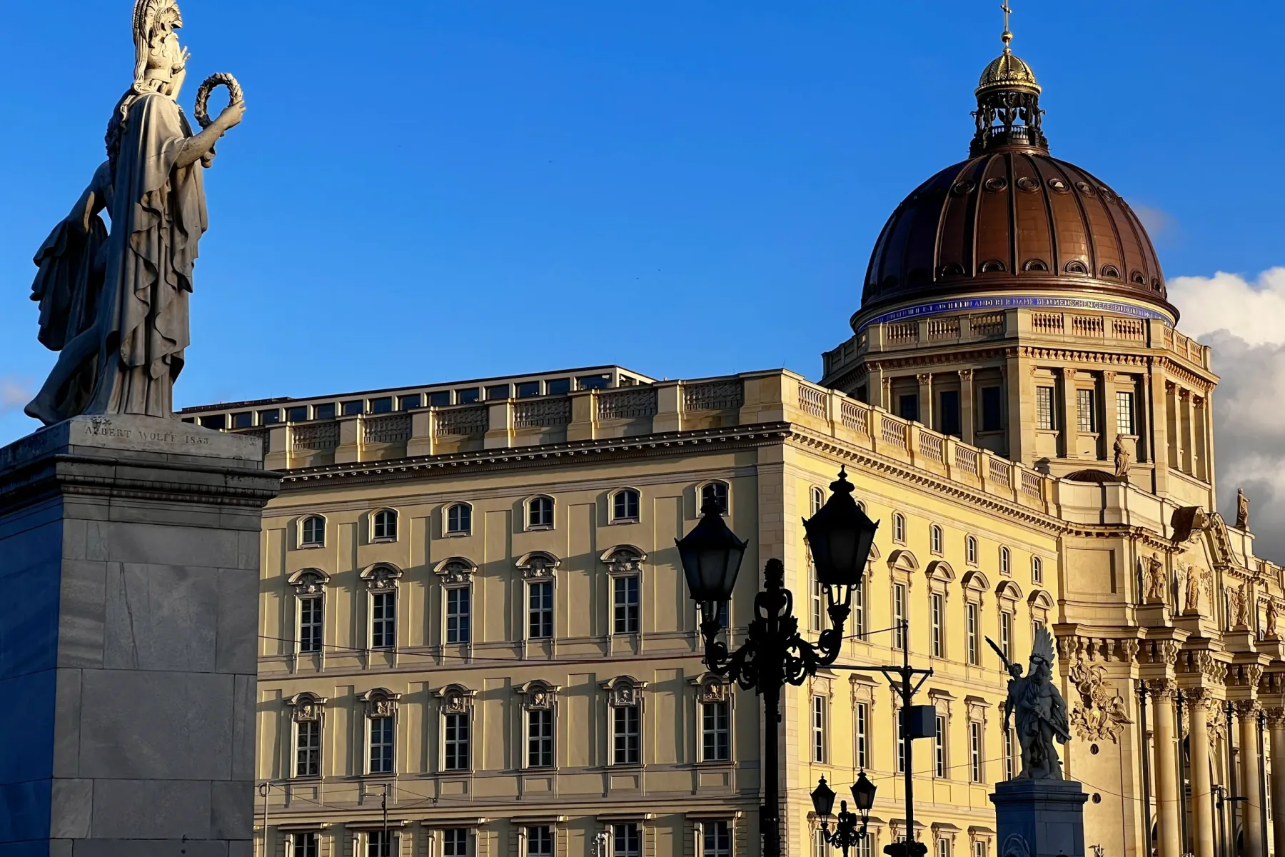 A classical stone statue of a female figure holding a wreath, with the dome and facade of the Humboldt Forum in Berlin beh...