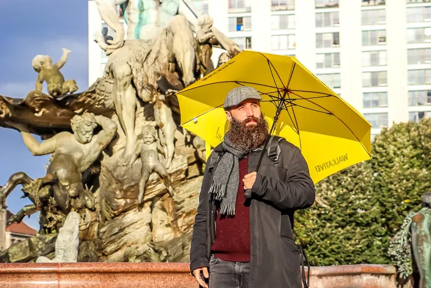 Bearded man in a hat holding a yellow Walkative umbrella, standing in front of the Neptune Fountain in Berlin.