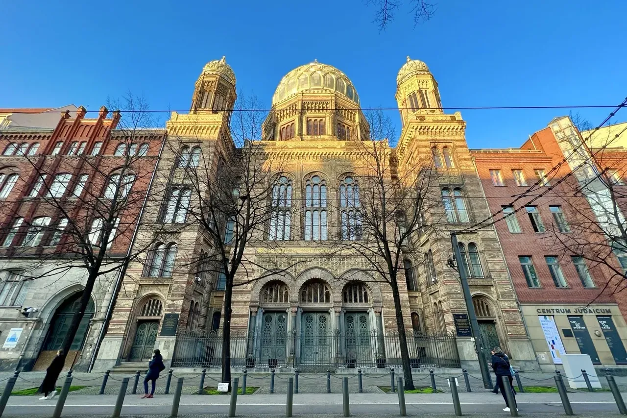 The ornate facade of the New Synagogue in Berlin on a sunny day, viewed from across the street through bare trees.