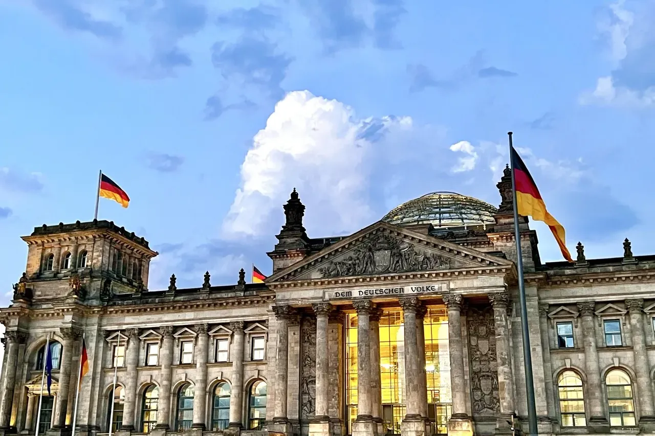 The Reichstag Building in Berlin at dusk, with German flags waving under a cloudy blue and white sky.