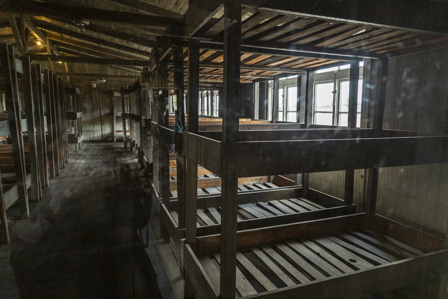 Innenansicht einer Holzbaracke mit mehrstöckigen Kojenbetten, lange Reihen im schwach beleuchteten Raum. Fenster sind im H...