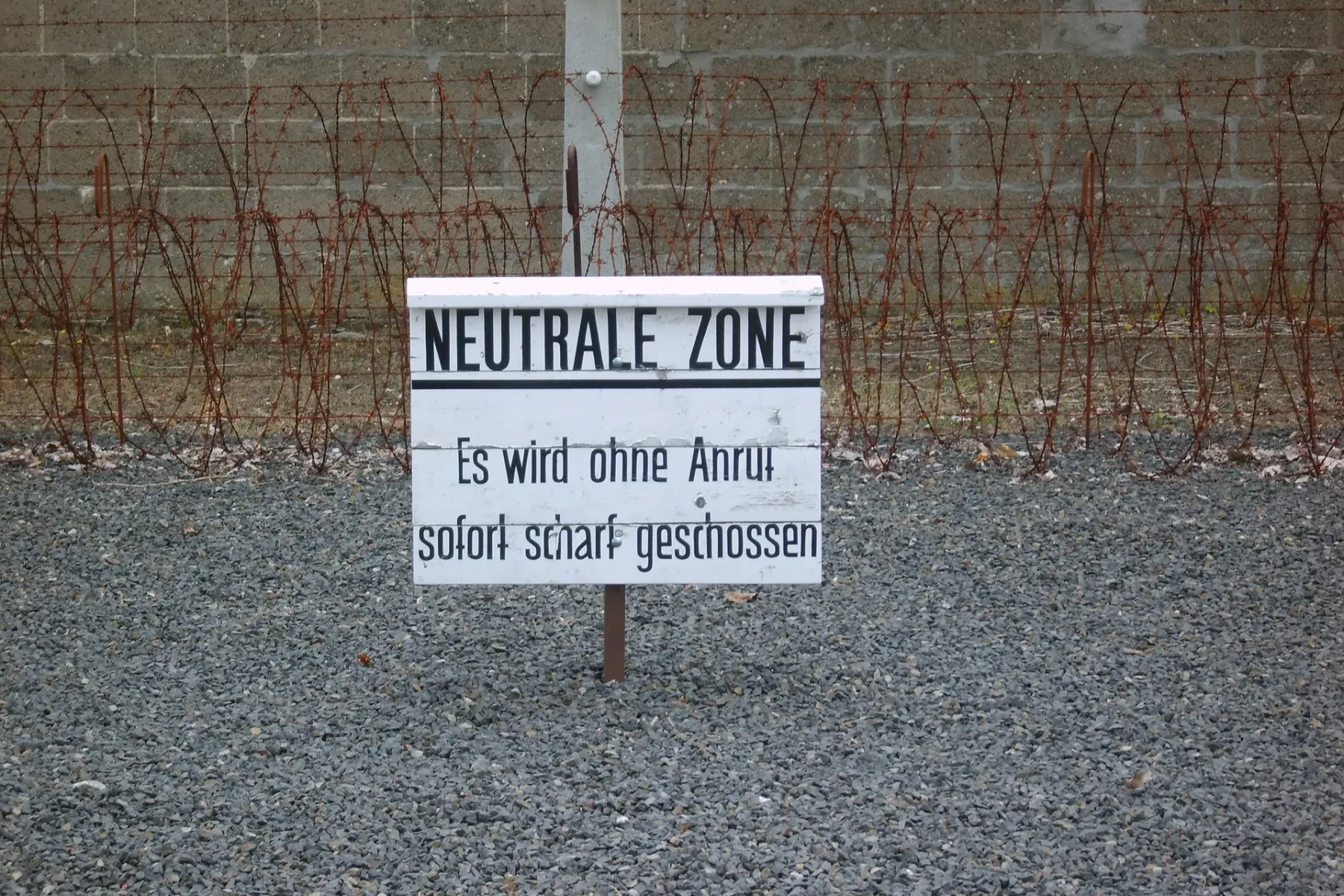 Weißes Schild mit "NEUTRALE ZONE" und Warntext auf Deutsch, vor Stacheldrahtzaun und Betonmauer auf Kiesboden.