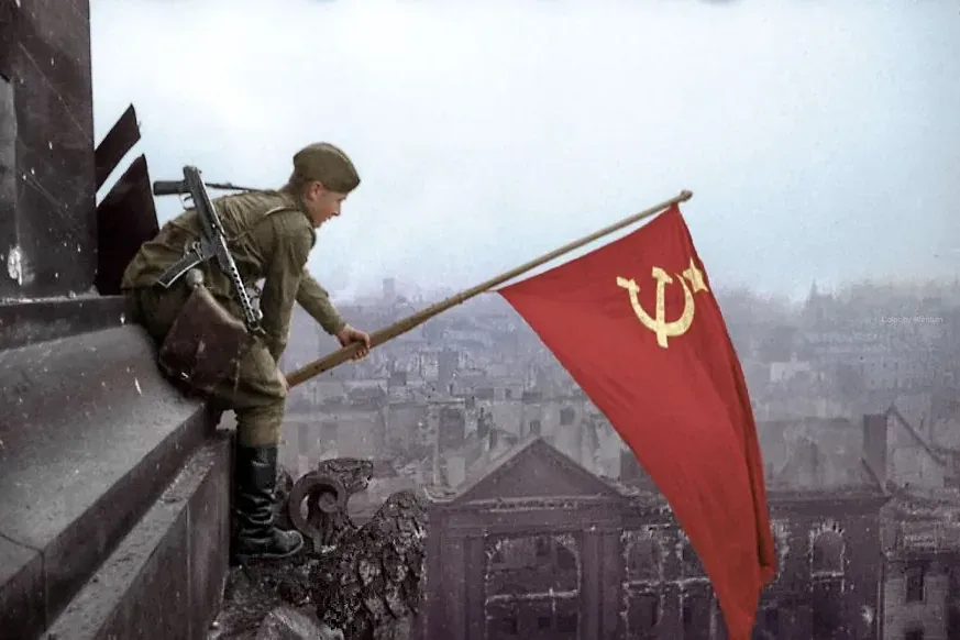 Soviet soldier planting a red flag with hammer and sickle on a destroyed building, city ruins in background.