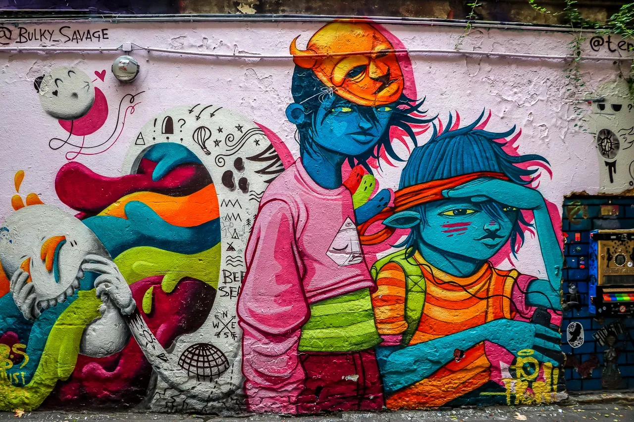 Farbenfrohes Streetart-Wandgemälde auf einer strukturierten hellrosa Wand mit stilisierten blauhäutigen Figuren und abstra...