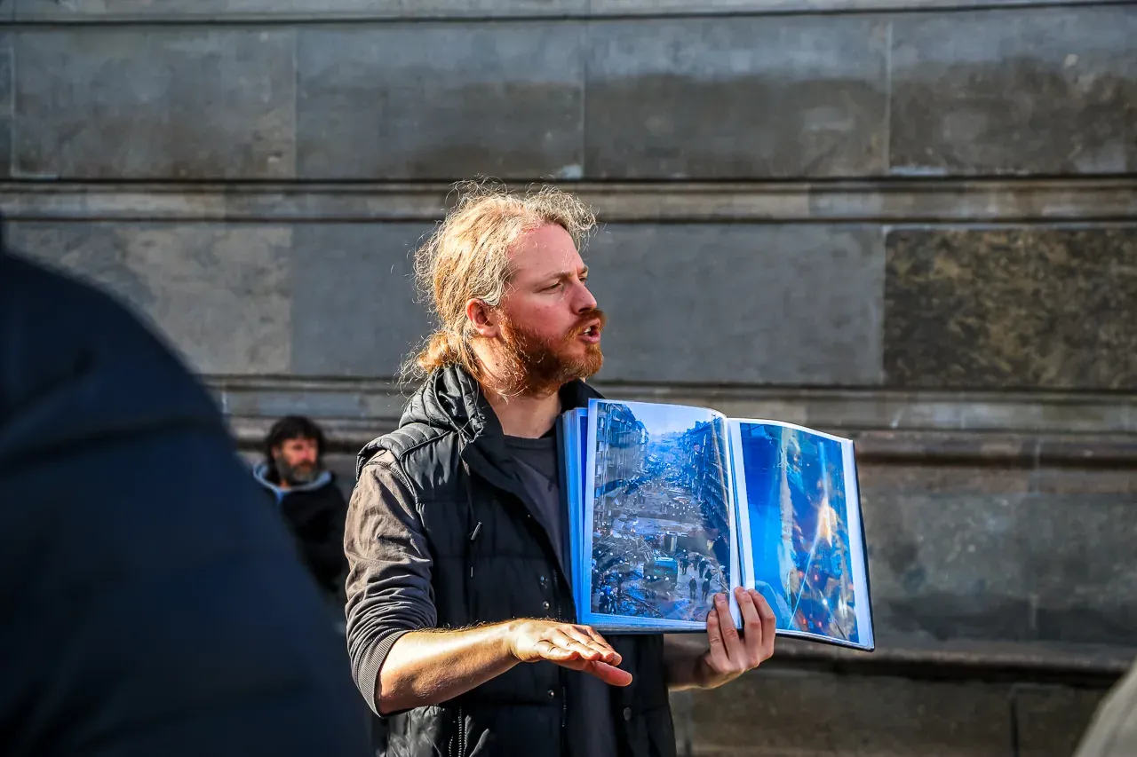 A tour guide in Berlin holds an open book showing a historical photo of a destroyed street while speaking to a group.