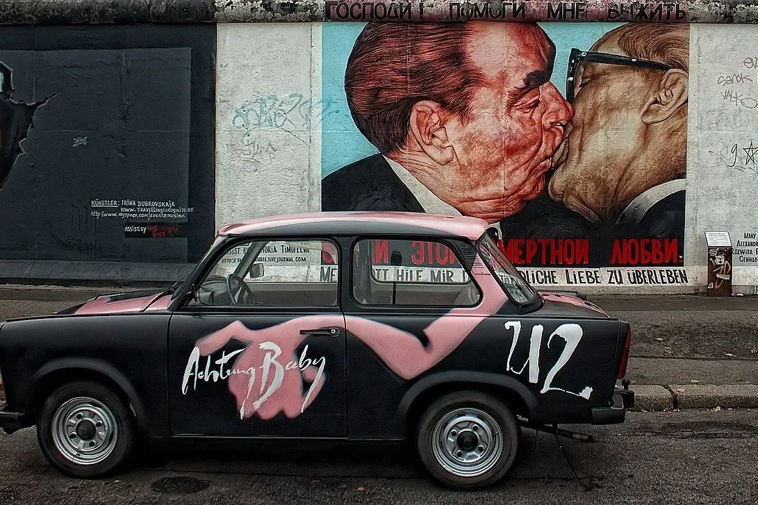 A black Trabant car parked in front of the 'Fraternal Kiss' mural on the East Side Gallery in Berlin.
