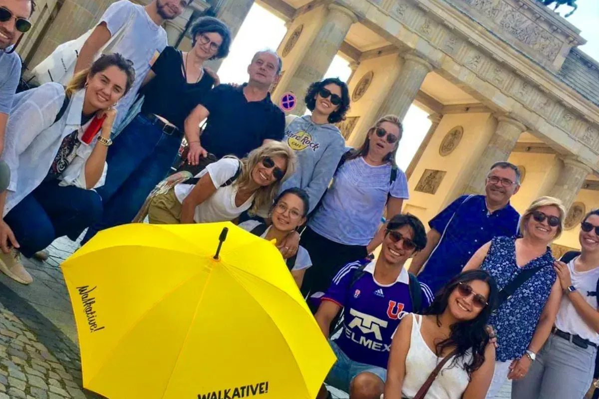 A diverse group of people posing with a yellow Walkative umbrella in front of the Brandenburg Gate in Berlin.