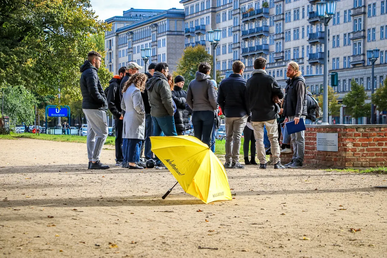 Un grupo de personas escuchando a un guía cerca de edificios de apartamentos en Berlín. Un paraguas amarillo de Walkative ...