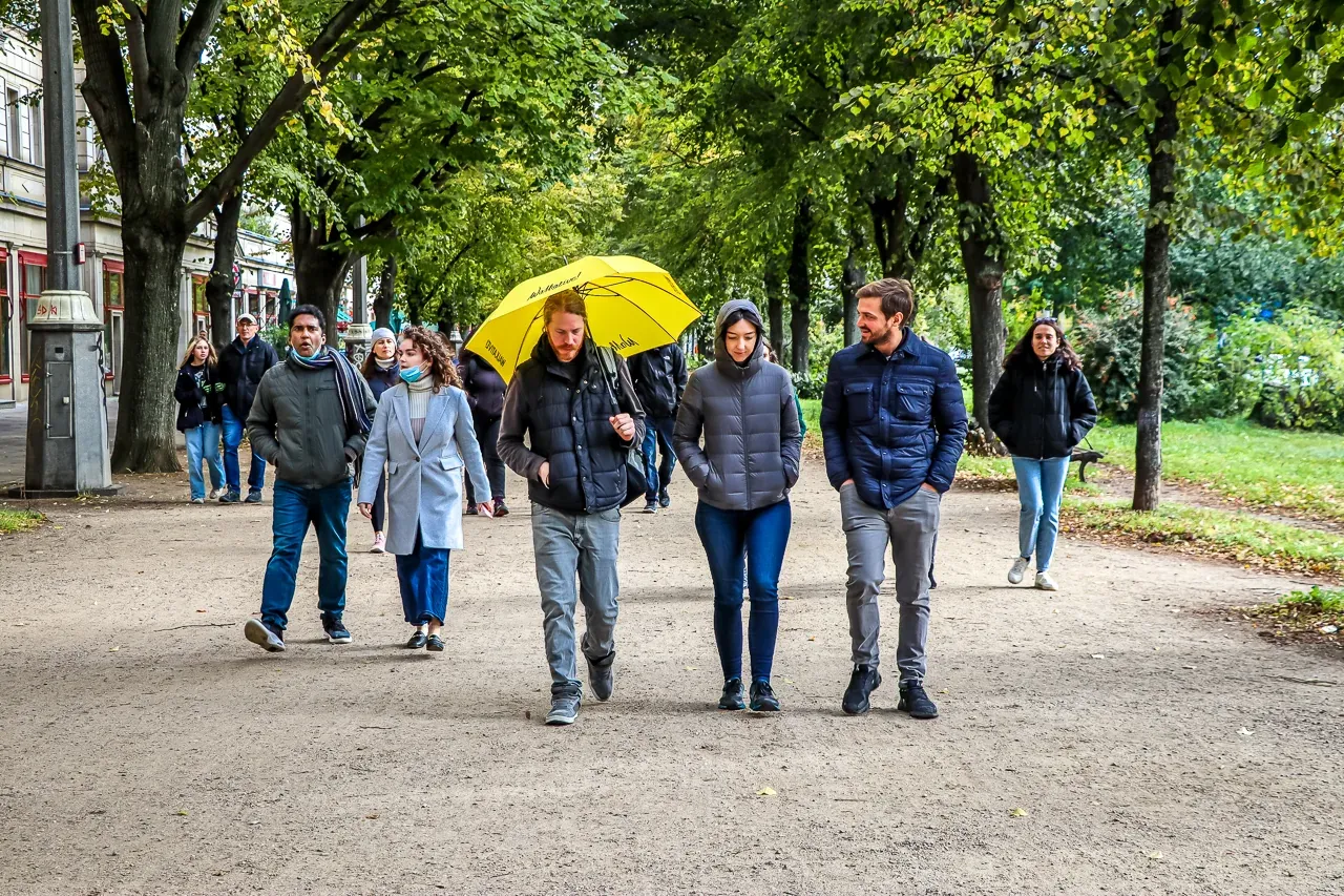 Group of people walking on a wide dirt path lined with mature trees. Buildings are visible on the left, one person holds a...