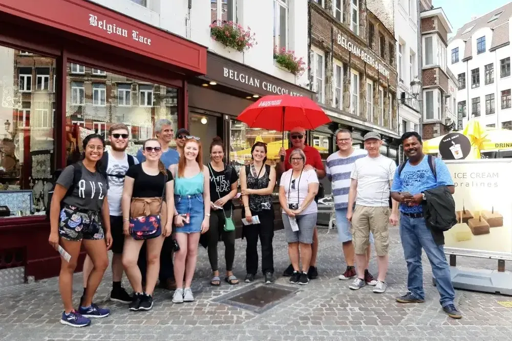 Un grupo diverso de turistas sonríe en una calle adoquinada frente a tiendas de encajes y chocolates belgas.