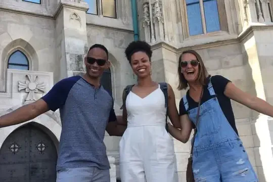Three smiling people stand arm-in-arm in front of the ornate, light-colored Matthias Church with Gothic windows.
