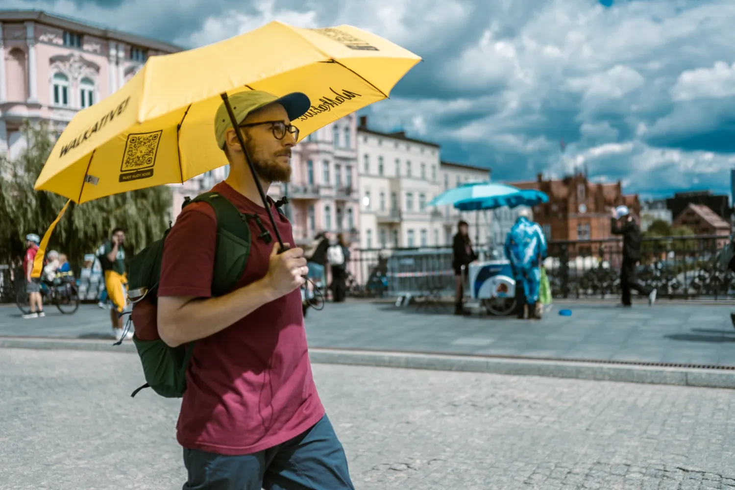 Przewodnik z brodą i w okularach trzyma duży żółty parasol Walkative, prowadząc wycieczkę po europejskim mieście.