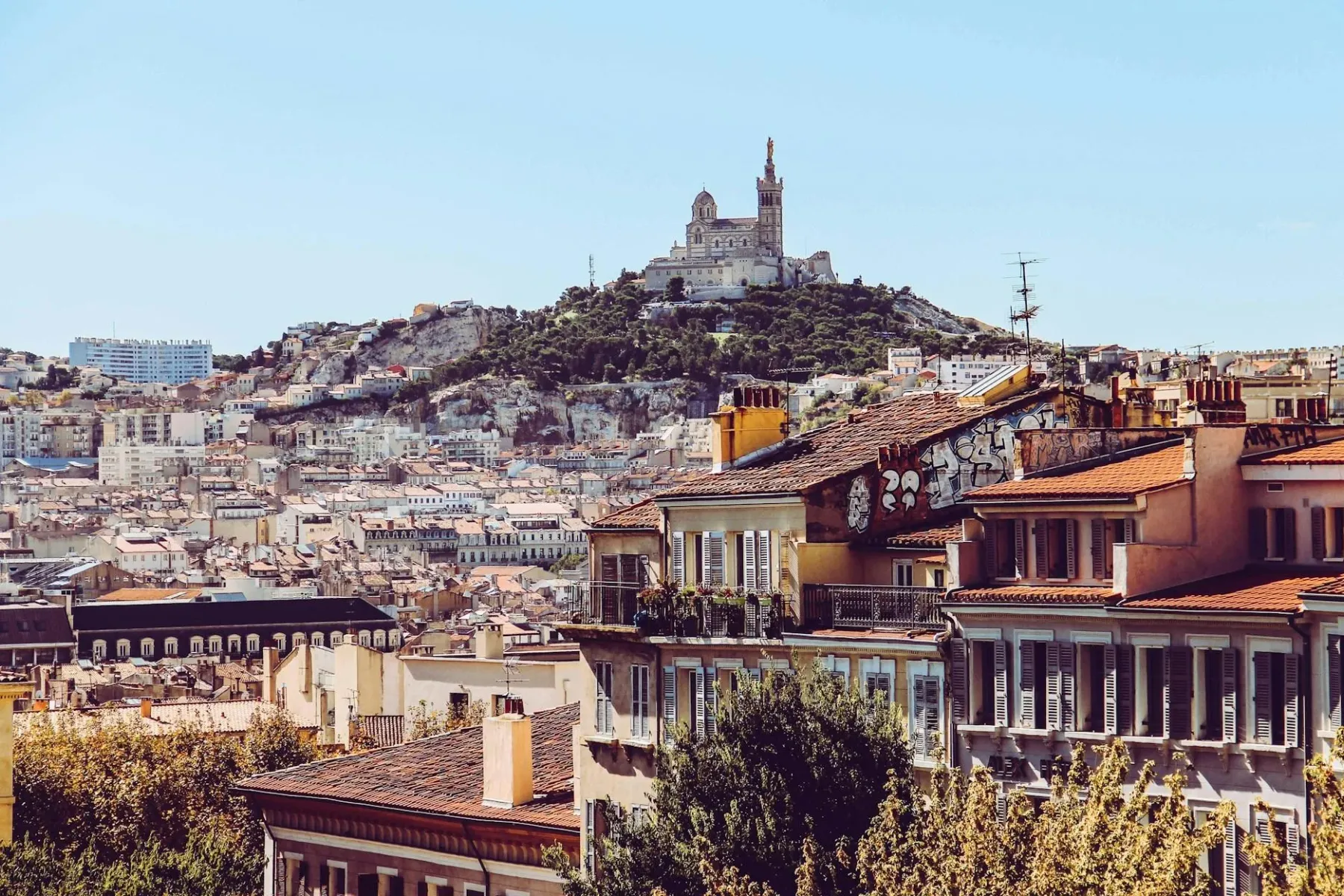 Vista panorámica de Marsella con la basílica Notre-Dame de la Garde en una colina, rodeada de edificios urbanos con tejado...