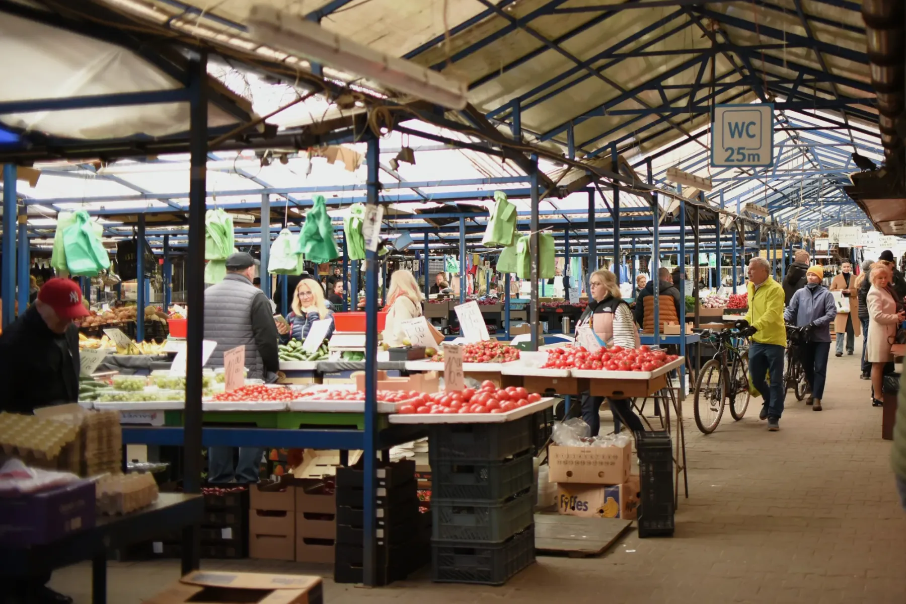People browse stalls laden with fresh red tomatoes and green vegetables inside a bustling indoor market with a translucent...