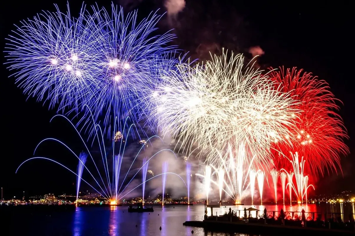 Blau-weiß-rote Feuerwerke explodieren nachts über einer Bucht. Eine Menschenmenge beobachtet vom Ufer; Stadtlichter spiege...
