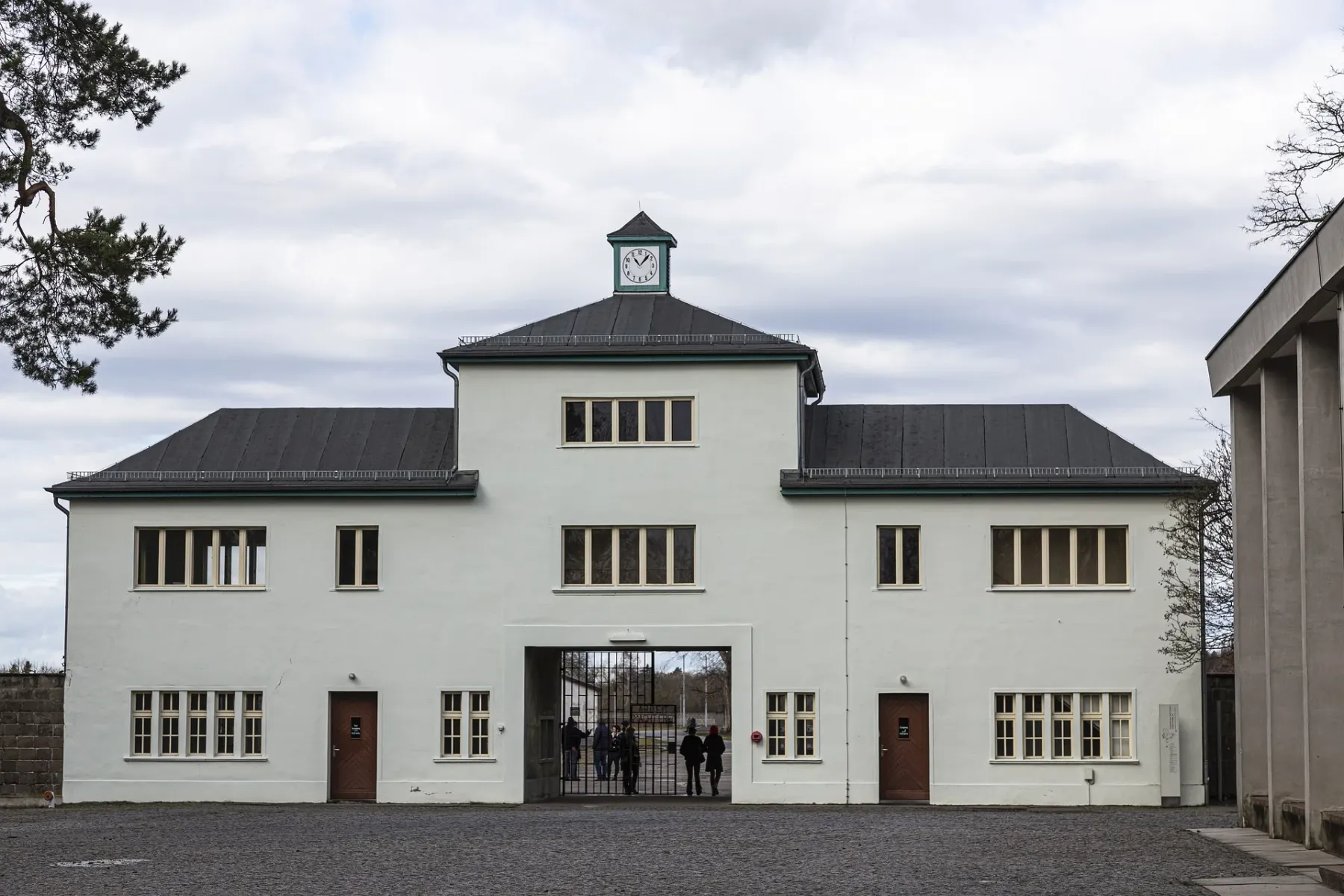 Weißes zweistöckiges Gebäude mit Uhrenturm und großem Eisentor. Menschen stehen am Eingang auf Kopfsteinpflaster unter bew...