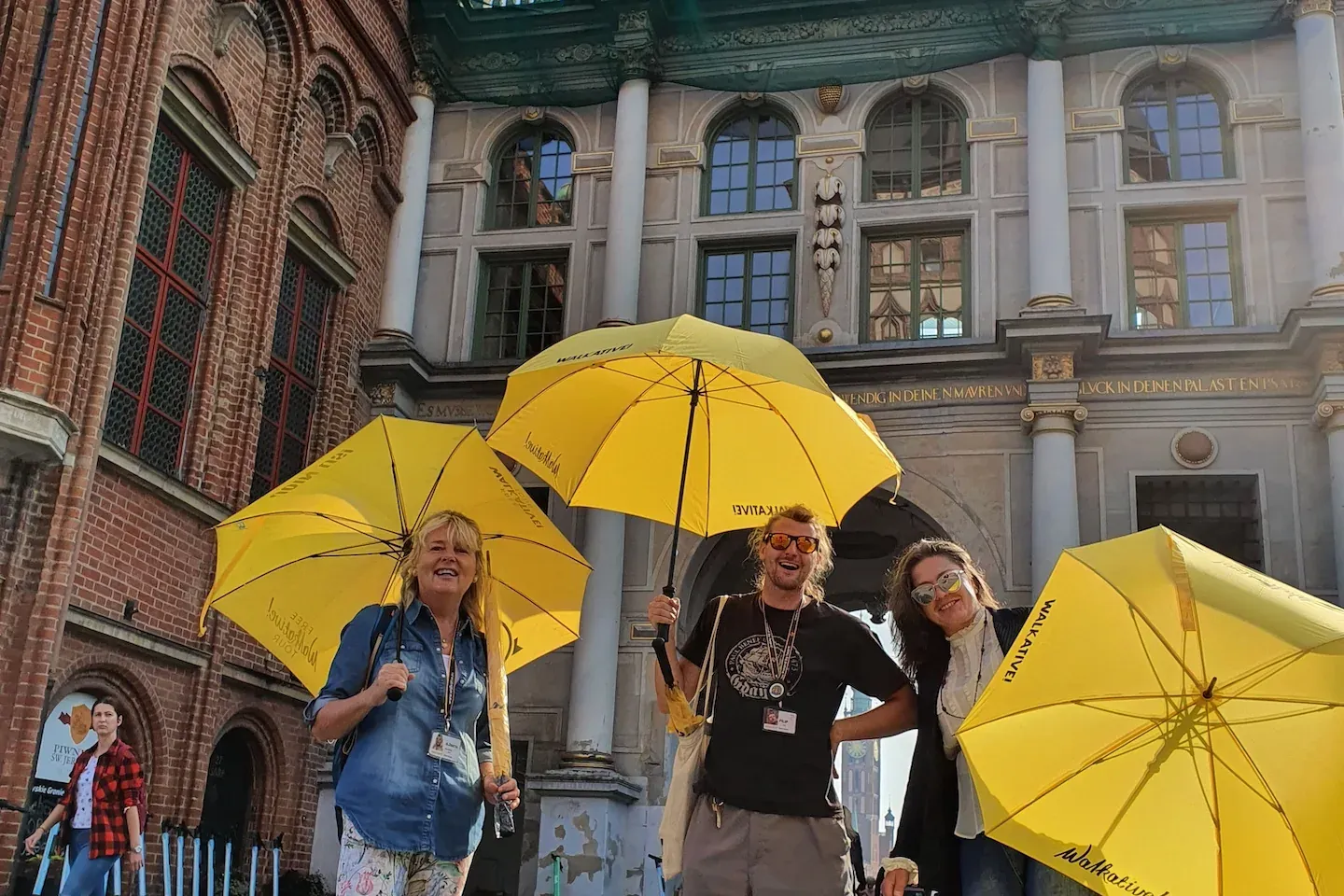 Trzy uśmiechnięte przewodniczki i przewodnik Walkative z żółtymi parasolami na brukowanej ulicy przed Zieloną Bramą w Gdań...
