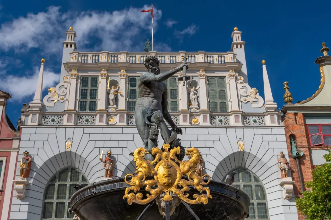 Bronzestatue des Neptun mit Dreizack auf einem Brunnen vor einem prächtigen, reich verzierten Gebäude bei blauem Himmel.