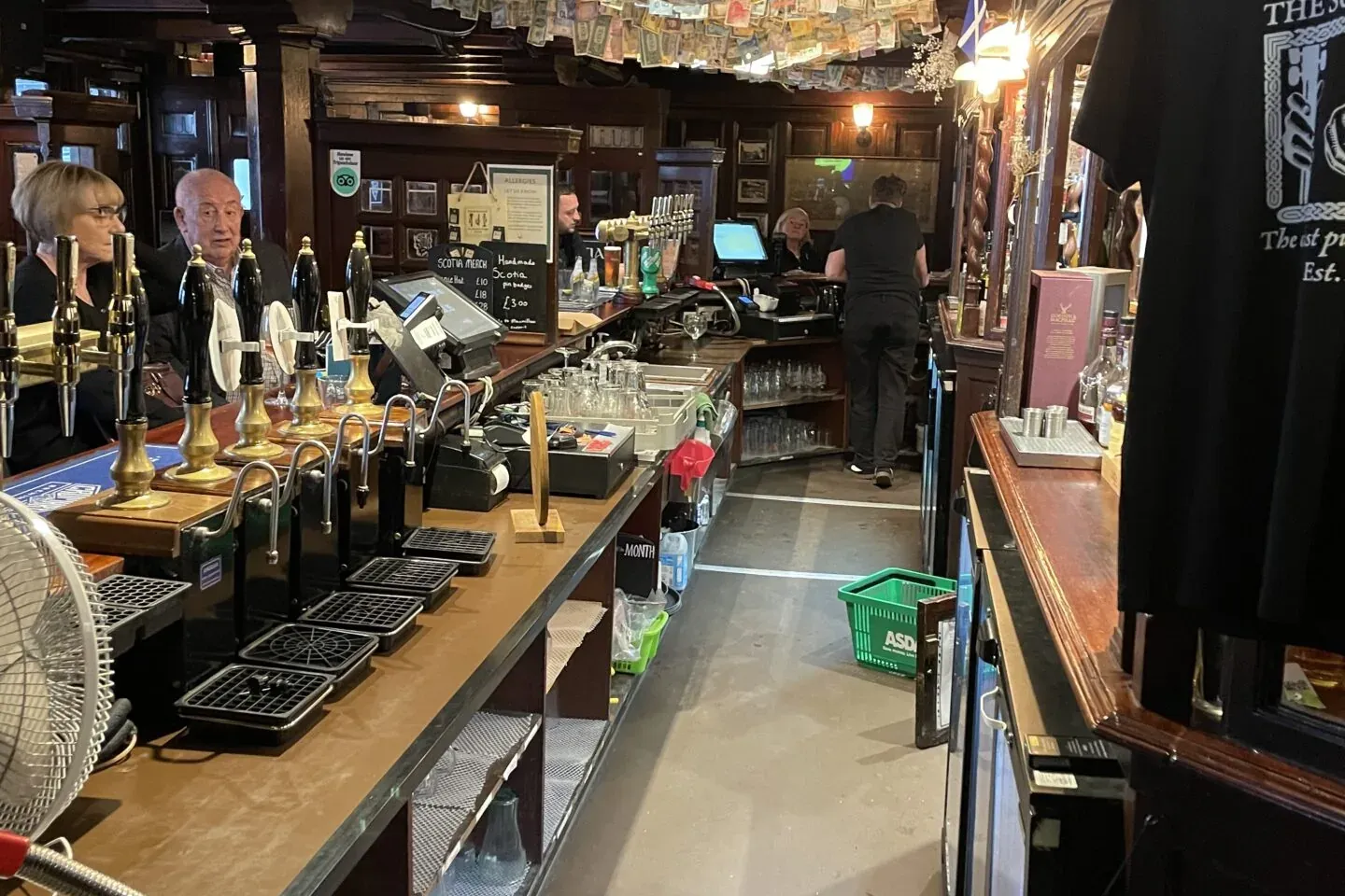 Interior de un pub tradicional de Glasgow: larga barra de madera, grifos de cerveza y billetes de varios países colgando d...