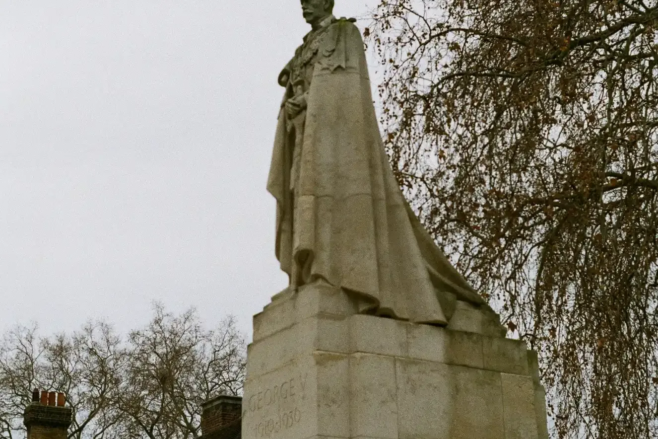 Estatua del rey Jorge V sobre un zócalo de piedra, con una paloma en la cabeza y árboles sin hojas al fondo.