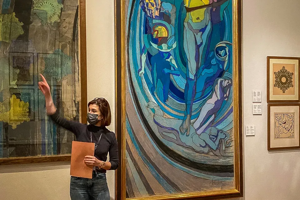 Eine Stadtführerin mit Gesichtsmaske steht in einer Kunstgalerie und zeigt auf ein großes, farbenfrohes Gemälde an der Wand.