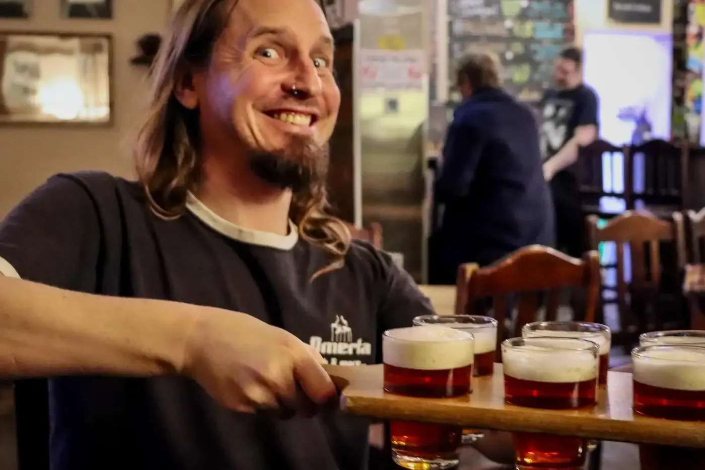 Ein Mann mit fröhlichem Gesichtsausdruck hält in einer Kneipe ein Holzbrett mit fünf kleinen Biergläsern.