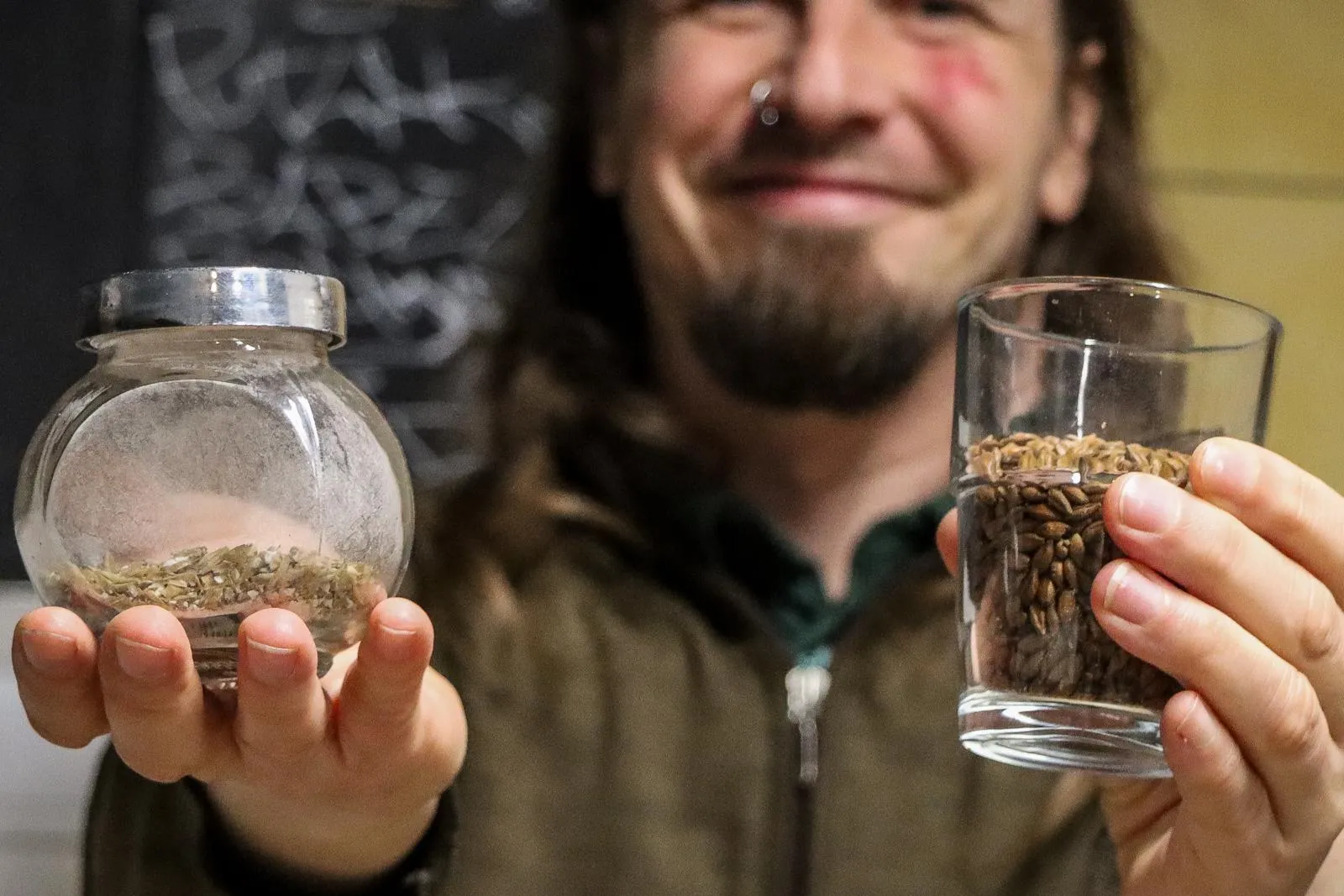 Ein Mann mit langen Haaren hält ein Glas mit Gerstenmalz und ein kleines Glas mit getrocknetem Hopfen in die Kamera.