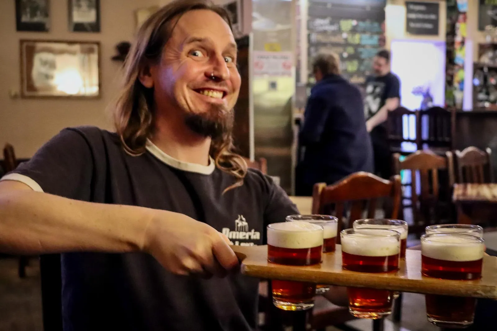 Ein lächelnder Mann mit langen Haaren und Nasenring hält in einem Pub ein Holzbrett mit sechs kleinen Gläsern Bier.