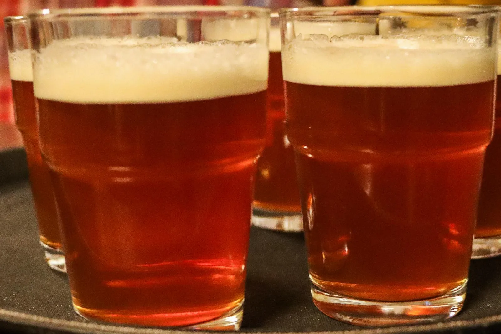 Mehrere Gläser bernsteinfarbenes Bier mit Schaumkronen stehen auf einem dunklen Serviertablett in einem Pub.