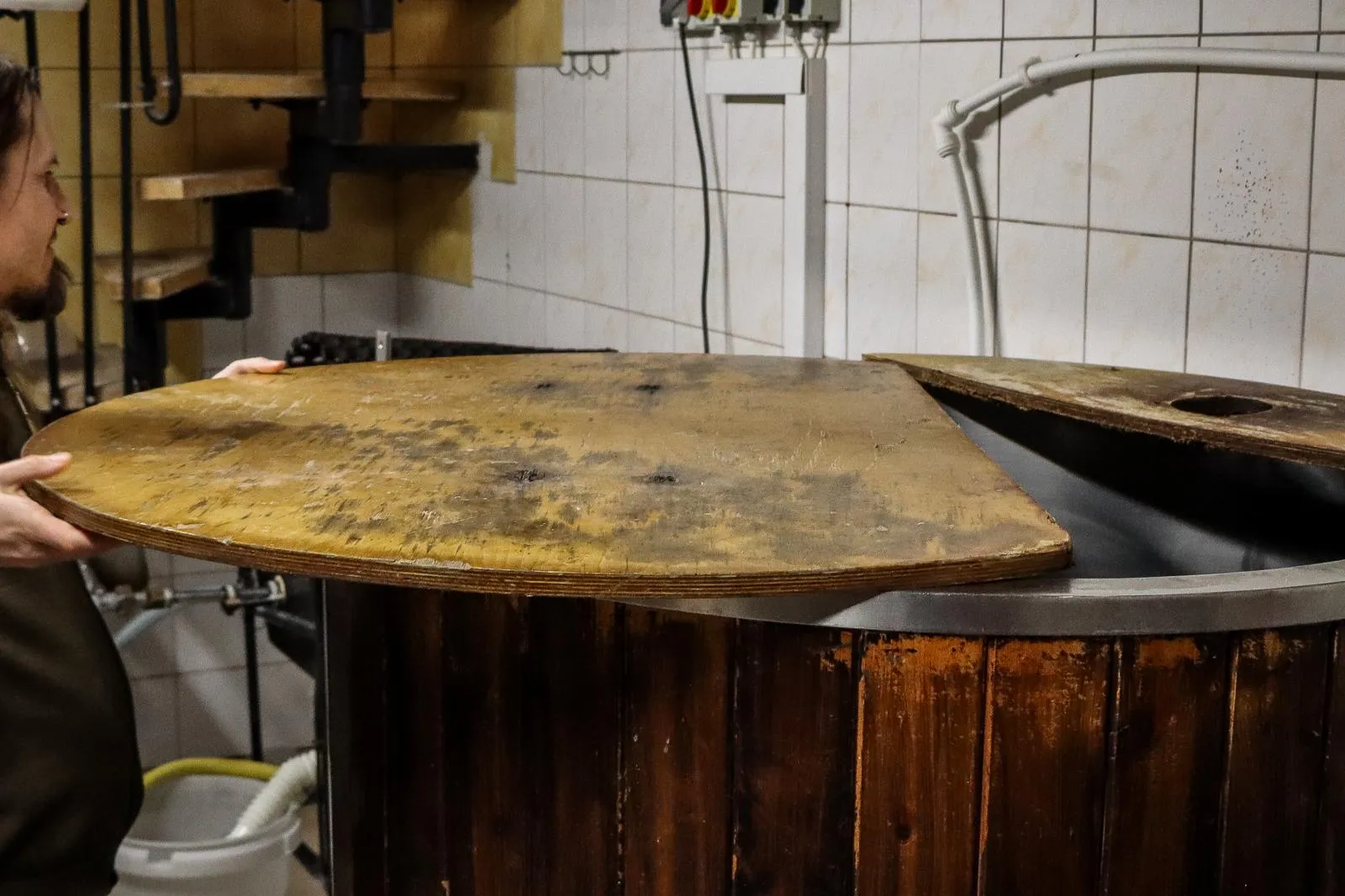 Ein Brauer öffnet den Holzdeckel eines großen Gärbottichs in einer kleinen Brauerei.