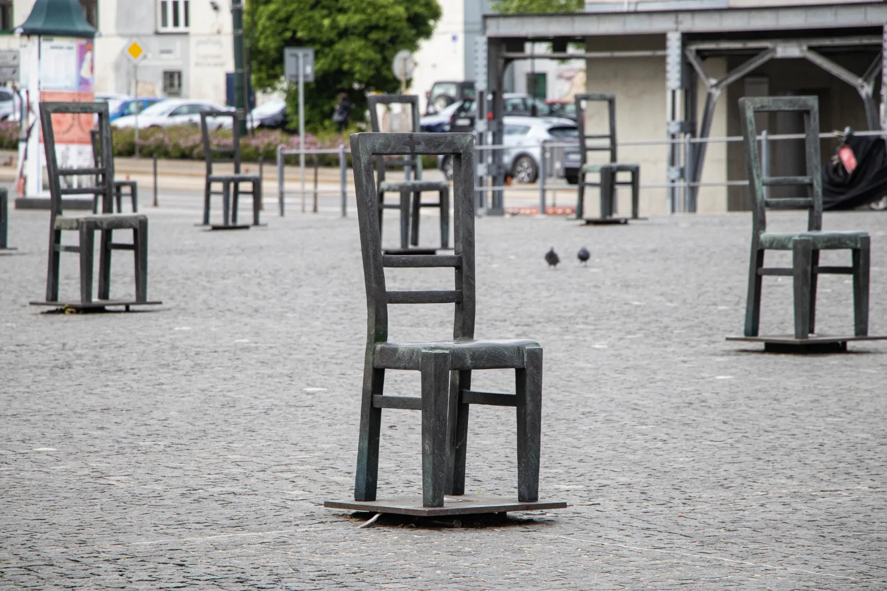 Esculturas de sillas de metal llenan una plaza adoquinada, con dos palomas visibles y edificios al fondo.