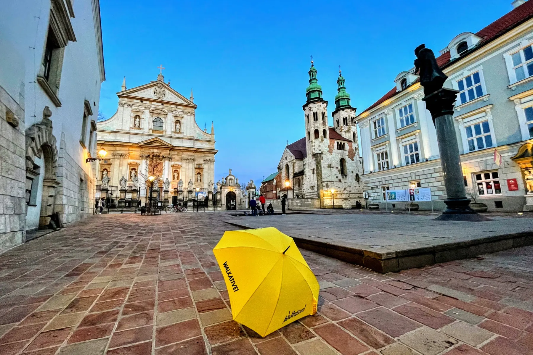 Ein gelber Walkative-Regenschirm auf dem Kopfsteinpflaster eines Platzes in Krakau, mit historischen Kirchen im Hintergrund.
