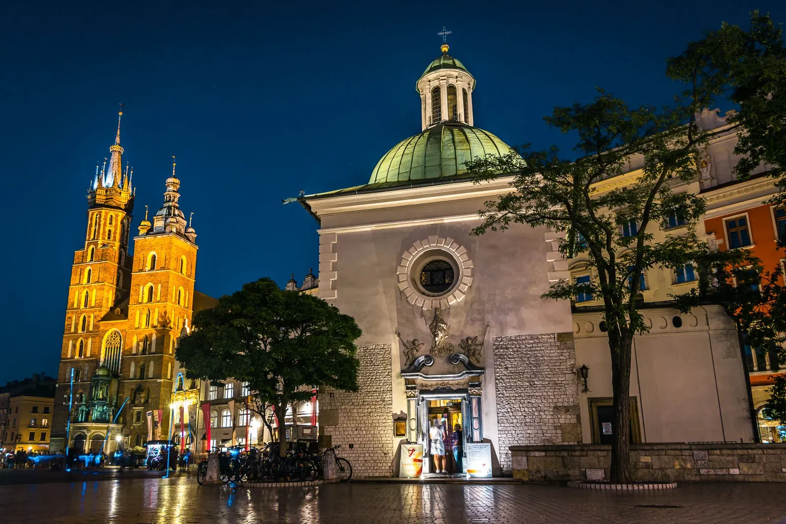 Marienbasilika und Adalbertkirche bei Nacht auf dem Krakauer Hauptmarkt beleuchtet, mit nassen Kopfsteinpflastern.