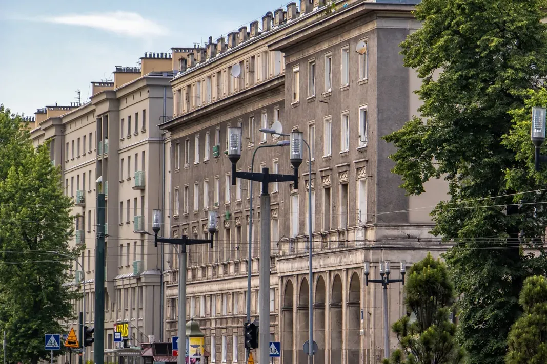 Eine breite Straße in Nowa Huta, Krakau, mit langen sozialistisch-realistischen Wohnblöcken, grünen Bäumen und blauem Himmel.
