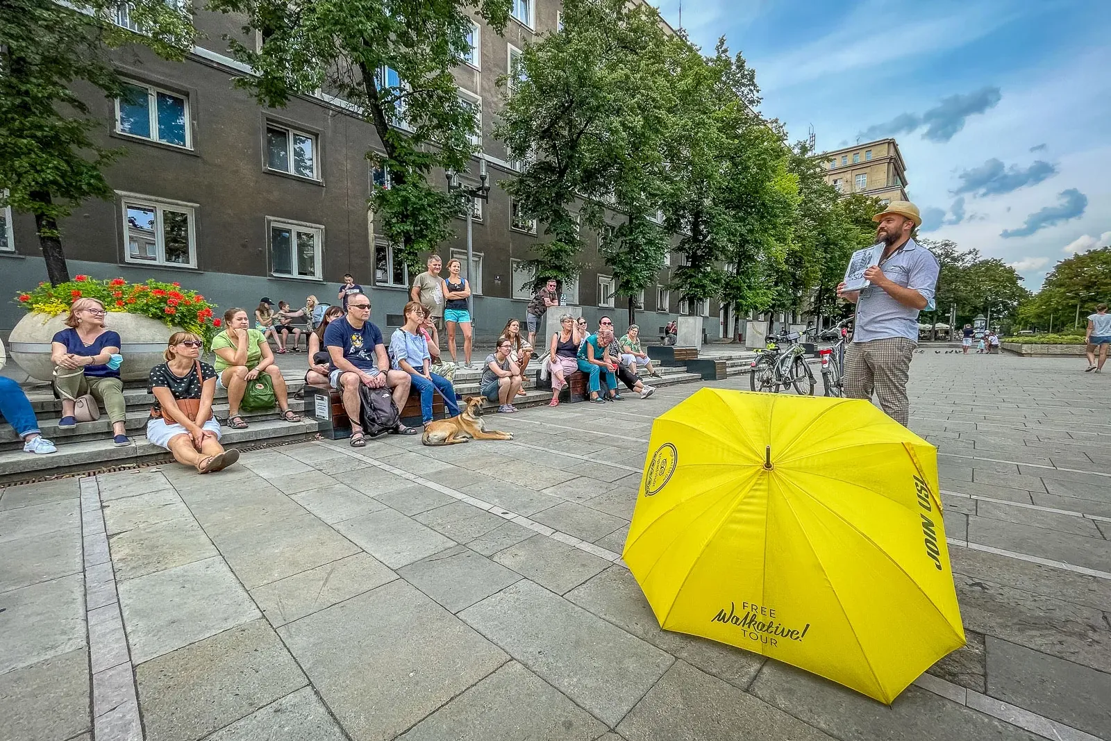 Ein Reiseführer spricht zu einer Gruppe von Leuten auf den Stufen eines Stadtplatzes, mit einem gelben Walkative-Schirm da...