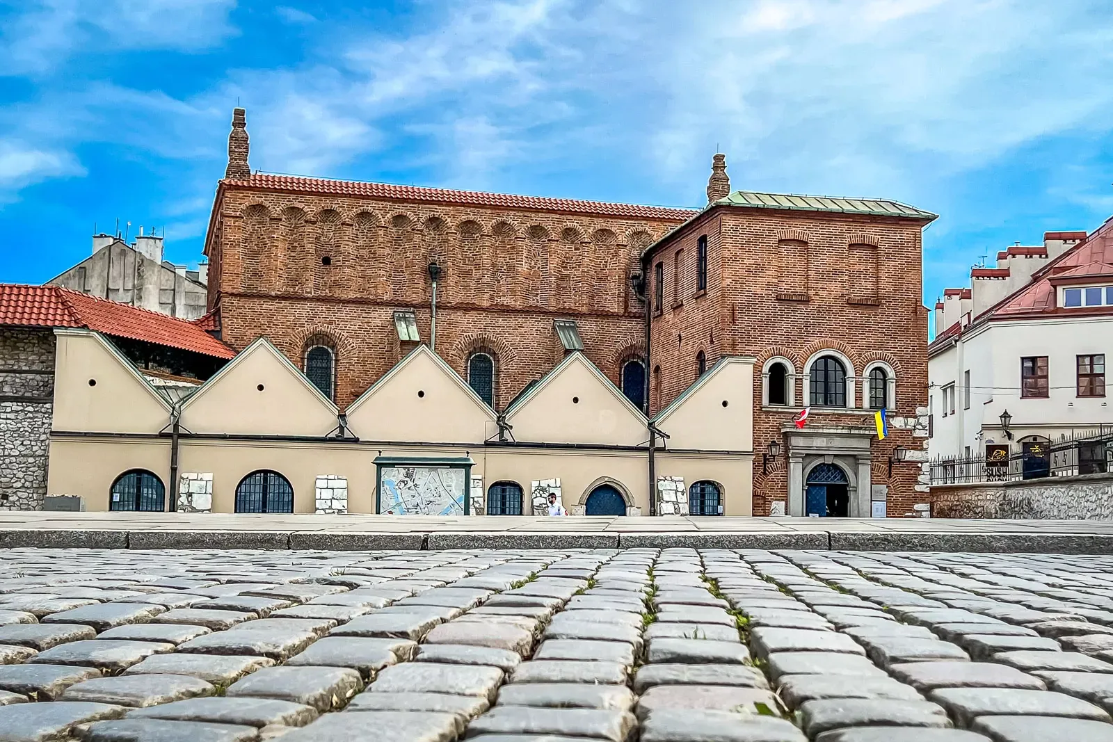 Alte Backsteinsynagoge mit Rundbogenfenstern, rotem Dach und Kopfsteinpflaster im Vordergrund unter blauem Himmel in Krakau.