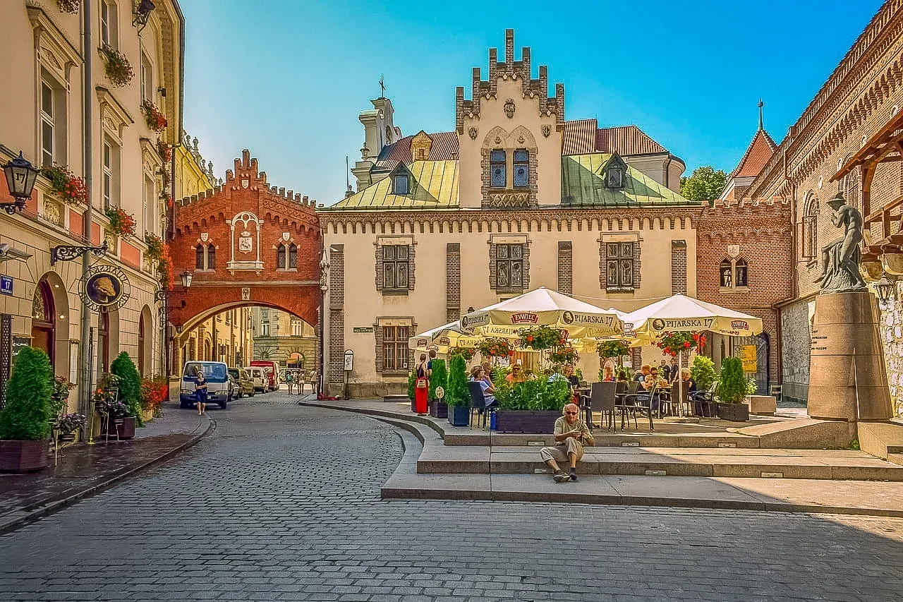 Kopfsteingepflasterte Straße in Krakau mit rotem Backsteinbogen, historischen Gebäuden und einem Straßencafé unter weißen ...