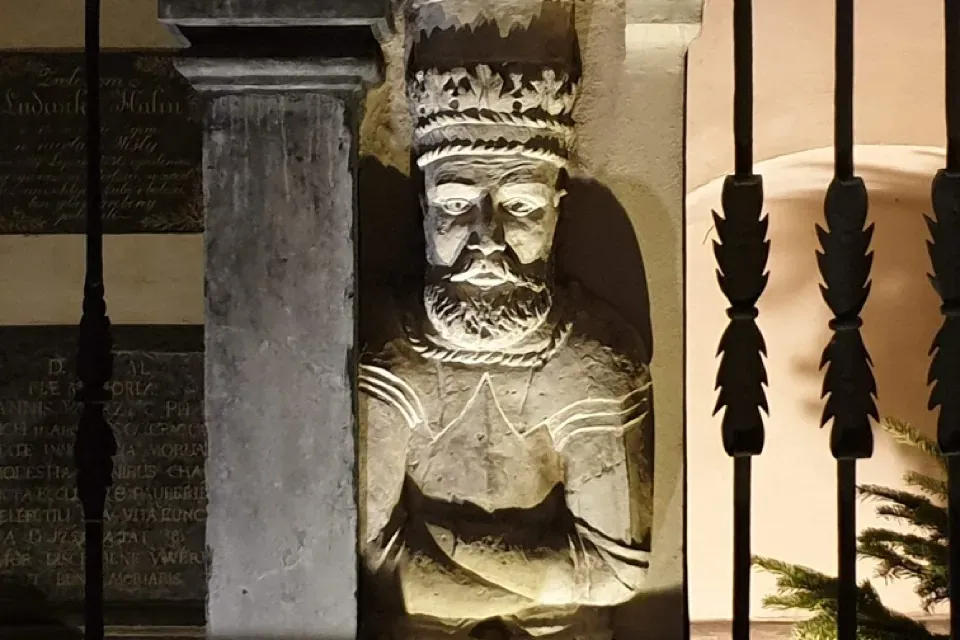 Eine Steinskulptur eines bärtigen Königs mit Krone, in einen Pfeiler eingelassen und nachts von unten beleuchtet.