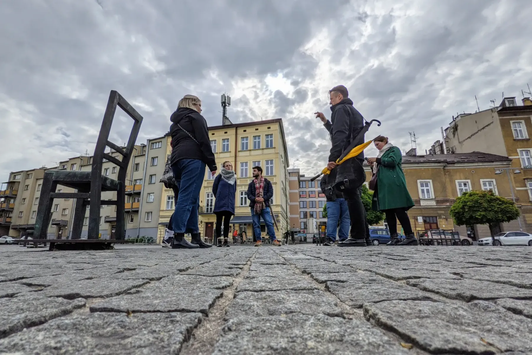 Un guía turístico habla con un pequeño grupo en una plaza adoquinada con grandes sillas de metal y edificios bajo un cielo...
