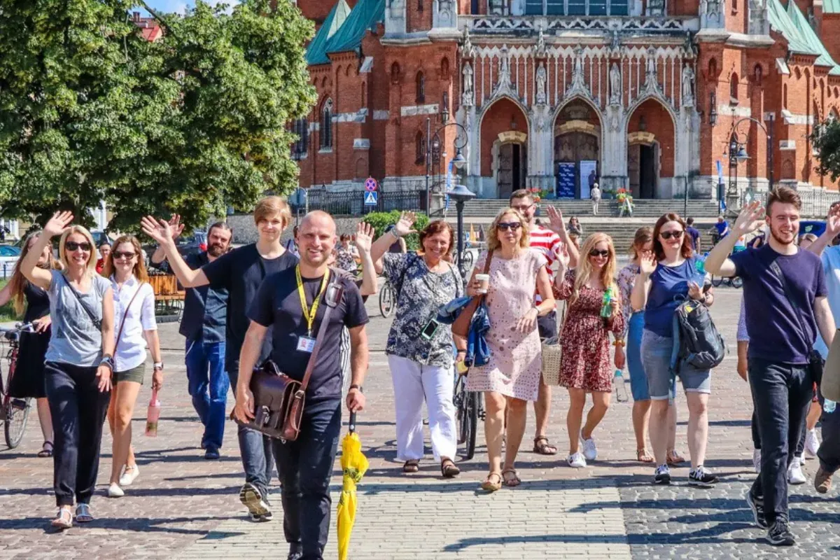 Ein Reiseführer mit gelbem Schirm führt eine Gruppe, die vor einer großen Backsteinkirche in Krakau in die Kamera winkt.